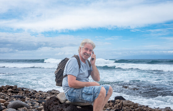 Cheerful elderly man with a white beard sitting on a rocky beach talking with his cell phone enjoying relax, retirement or travel. Horizon over water
