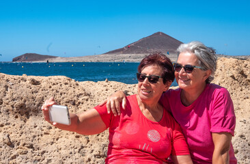 Happy relaxed couple of elderly women enjoying summer vacation at sea, sitting using phone for...
