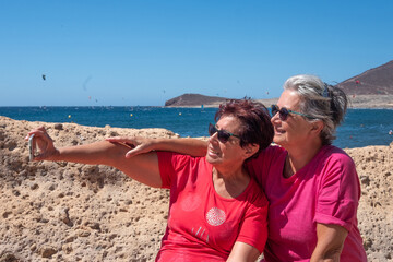 Happy relaxed couple of elderly women enjoying summer vacation at sea, sitting using phone for...
