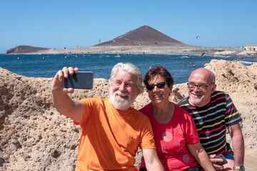 Cheerful group of seniors enjoying a summer vacation at sea, sitting using phone for selfie in a...