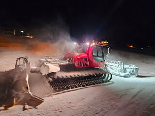 A snow plow groomer is driving down a snowy road, red and white and has a large scoop on the front....