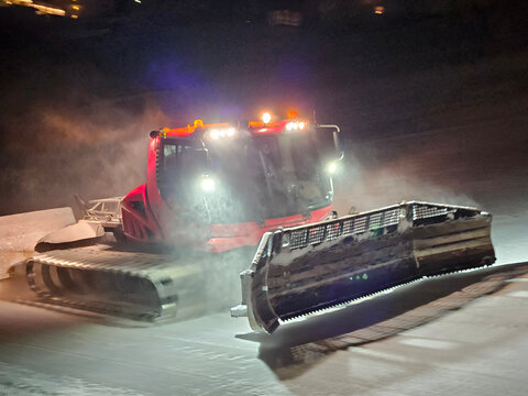 A snow plow is driving down a snowy road. The plow is red and has a black and white stripe. Groomer ratrak is clear that it is working hard to clear the road on ski resort