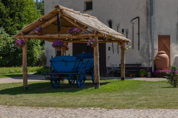 Rustic wooden gazebo with hanging flower pots and vintage blue cart on green grass in countryside yard near old farmhouse on sunny summer day. Rural lifestyle, outdoor decor and tradition
