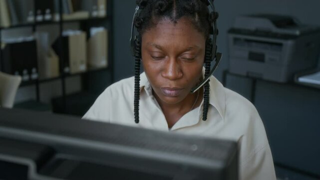 Close-up view of Black young female customer support operator with microphone headset clarifying clients issue and using computer at workplace