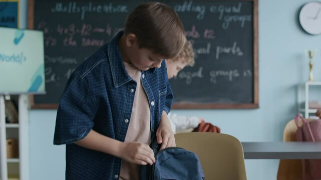 Medium shot of Caucasian schoolboy standing by his desk as taking notebooks and pencil case off his backpack, preparing for classroom lesson, with his classmate and board in background