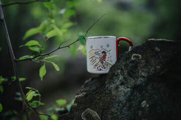 Colorful travel mug with an outdoor design placed on a rock amidst greenery, emphasizing nature and...