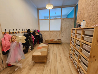 A little girl in a pink dress is standing in a room with a rack of coats and a bench