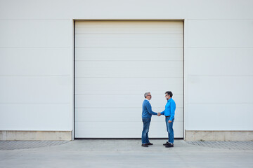 Fototapeta premium Two Businessmen Shaking Hands in Front of a White Industrial Building