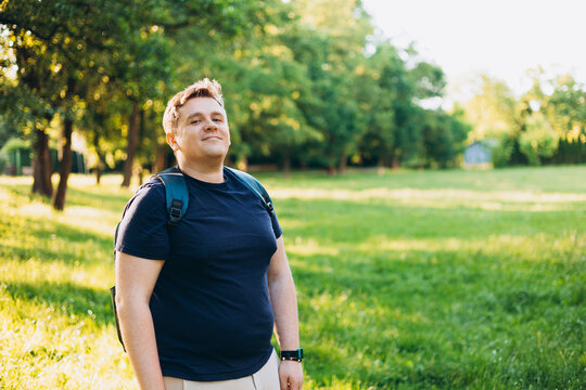 Happy 30s blonde man with backpack standing in the countryside. Traveler. Summer time. Portrait of happy young man in casual clothing standing outdoor