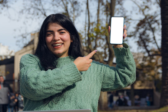 Venezuelan woman outdoors smiling, looking at the camera and showing the blank screen of her phone