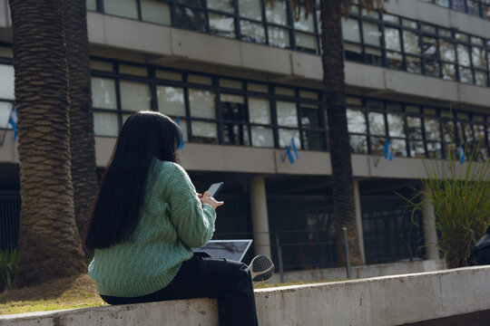 woman with her back turned sitting in the square using her cell phone and computer.