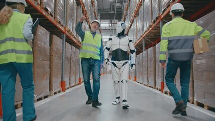Supervisor in reflective vest pointing upward while walking with humanoid robot through warehouse aisle. Two workers in safety gear moving past. Carrying box and checking tablet during active workday. - Powered by Adobe