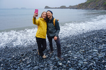 Couple enjoys Turkish coastline and taking selfie on beach in Winter. walking on stones, watching sea waves, relaxing together, slow romantic moment near calm blue water under soft winter light.