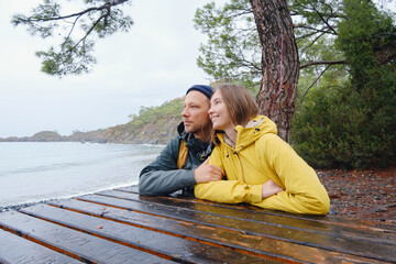 Couple enjoys Turkish coastline. Sitting on stones, watching sea waves, relaxing together, slow romantic moment near calm blue water under soft winter light.