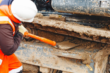 Construction worker  in bright safety gear working hard using shovel to clean tracks of his heavy...