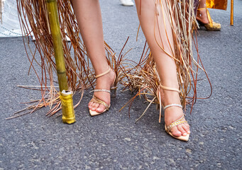 Close-up of a woman's legs wearing woven high-heeled sandals, with decorative fringe hanging from her outfit. Stylish detail from a festive street event
