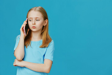 Sad girl in a blue shirt talking on a smartphone, against a bright blue background, expressing...