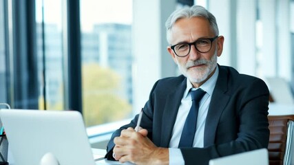 Confident senior businessman with gray hair and beard wearing suit and glasses sitting modern office desk with laptop, holding pen, smiling, looking camera, professional atmosphere, natural light - Powered by Adobe