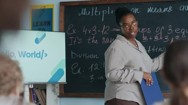 Medium shot of Black female teacher pointing at rules written with chalk on board while explaining multiplication to young students in modern classroom