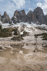 Spring snowmelt and mountain reflection at foot of Geisler peaks in the Dolomites, with cloudy sky and alpine forest landscape.