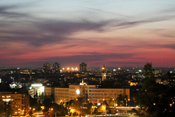 Twilight view of Novi Sad, Serbia, with city lights glowing beneath a dramatic sky and visible landmarks like church towers and residential buildings.