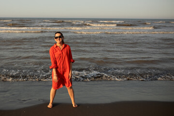 Happy woman in a red dress enjoys a peaceful beach walk at sunset, radiating joy and freedom during a relaxing seaside vacation. Travel moments full of emotion