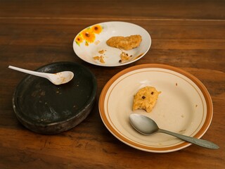 Three plates with leftover food, including a half-eaten fried tempeh, placed on a wooden table with a spoon and a stone mortar