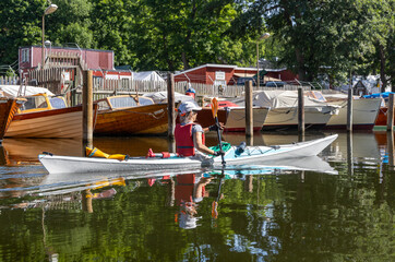 Woman kayaking past classic wooden boats and red boathouses in a quiet marina in central Stockholm.