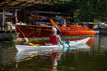 Woman kayaking past a gleaming wooden cabin cruiser moored beside a boat shed in central Stockholm.
