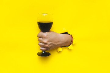 A man's hand emerges through a torn hole in yellow paper with a glass of red wine. The concept of...