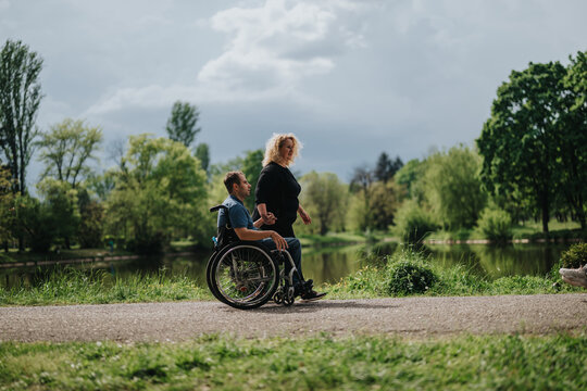 A person in a wheelchair and their companion are strolling along a path in a scenic park by a calm lake, surrounded by lush greenery and cloudy skies. - Powered by Adobe