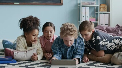 Four diverse schoolchildren in casualwear lying on printed black-and-white rug on floor in modern classroom, using digital tablet and chatting, resting before or between lessons - Powered by Adobe