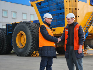 Two men shake hands in front of a large construction vehicle. One man is wearing an orange safety...