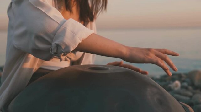 Woman playing handpan instrument by the sea during peaceful sunset moment