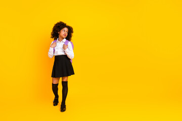 Portrait of a happy schoolgirl in uniform and backpack standing against a yellow background,...