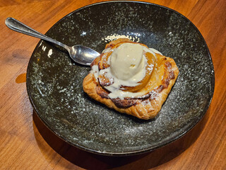 A dessert apple pie with a scoop of ice cream on top. The dessert is in a black bowl. The bowl is...