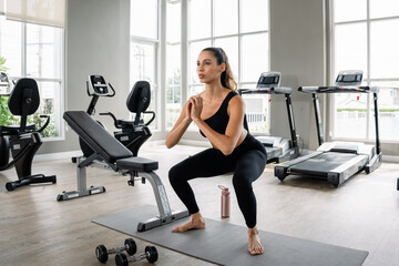 Fototapeta premium A woman is doing squats on a mat in a gym. There are several pieces of gym equipment in the background, including a treadmill, a stationary bike, and a bench