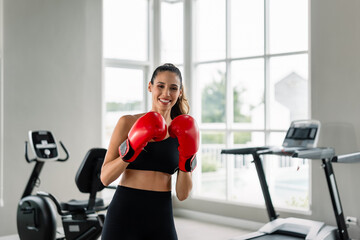 A woman is wearing a red boxing glove and standing in front of a stationary bike. She is smiling...