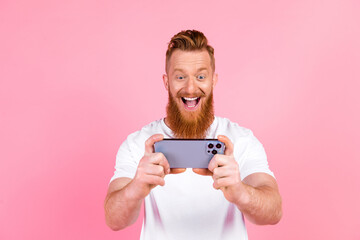 Excited red-haired man with a beard using a smartphone against a vibrant pink background in casual...