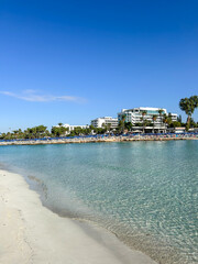Peaceful beach with soft white sand and shallow, clear turquoise water in front of a modern seaside resort under a bright blue sky, ideal for vacation and relaxation themes.