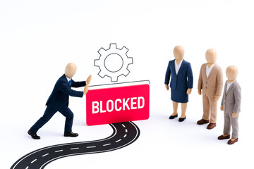 Miniature scene depicting person business suit pushing against large BLOCKED sign with gear icon above, symbolizing concept of obstacle or restriction professional or technological environment