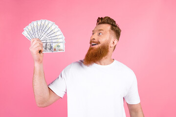 Young red haired man holding fan of dollar bills and smiling on pink background, symbolizing...