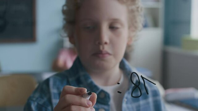 Smart Caucasian boy in blue plaid shirt standing by glass board with thoughtful expression on his face as solving division problem during Math lesson in modern, bright classroom