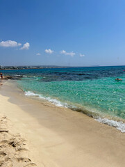 Golden sand and crystal-clear turquoise water on a peaceful beach in Cyprus, with distant buildings and a bright blue sky.