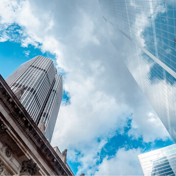 Financial Architecture, City of London. The landmark Tower 42 building reflected on a modern Bishopsgate skyscraper blending into a cloudy blue sky.