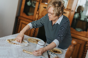 An elderly woman arranges food at a dining table in a cozy home, displaying domestic routines. The...