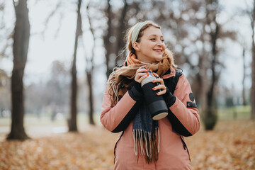 A cheerful woman, dressed warmly in a jacket and scarf, holds a thermos in an autumn woodland...