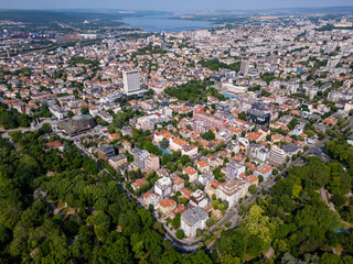 Aerial panoramic view of Varna, Bulgaria, showcasing the urban landscape with red rooftops, green parks, and the Black Sea in the distance under a clear blue sky