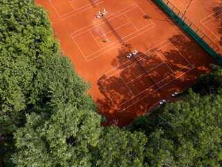Aerial view of red clay tennis courts surrounded by lush green trees. Empty courts with shadows and equipment create a peaceful and organized sports atmosphere