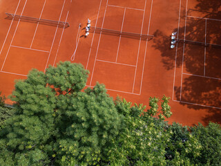 Aerial view of red clay tennis courts surrounded by lush green trees. Empty courts with shadows and equipment create a peaceful and organized sports atmosphere